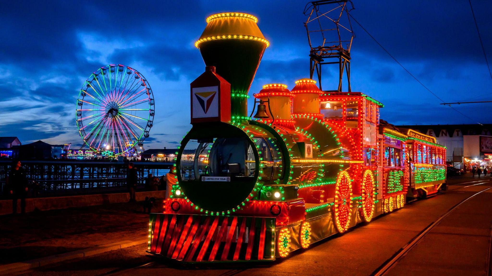 An illuminated Blackpool tram shaped like a steam locomotive on the promenade at dusk, with the Central Pier big wheel lit up behind